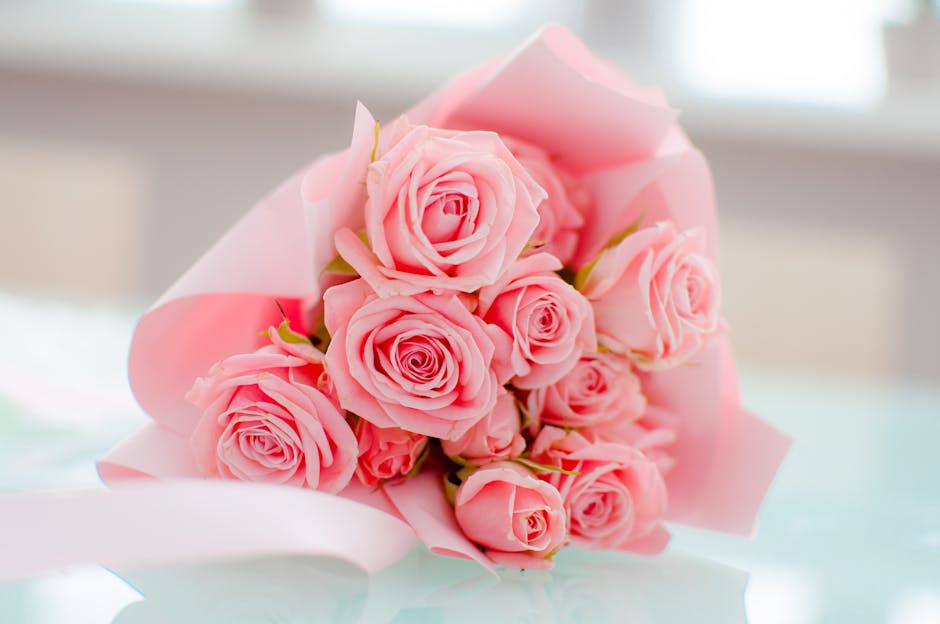 A bouquet of fresh pink roses with soft, velvety petals arranged in a tight, lush cluster. The roses are wrapped in delicate pink tissue paper, slightly fanned out at the edges, and are displayed on a