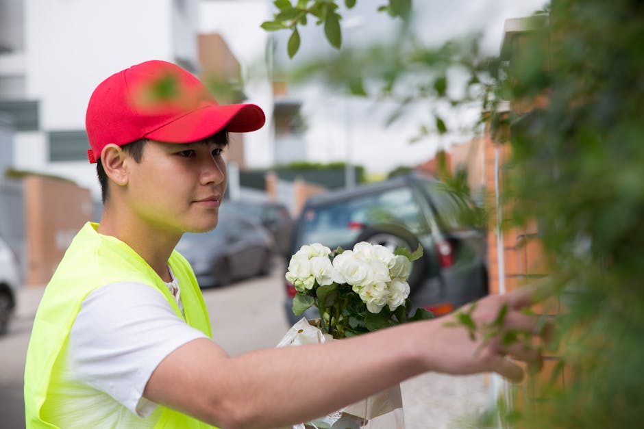 A delivery person wearing a red cap and a bright yellow safety vest is holding a bouquet of white flowers with green foliage, preparing to deliver it in a residential area. The person is standing outd